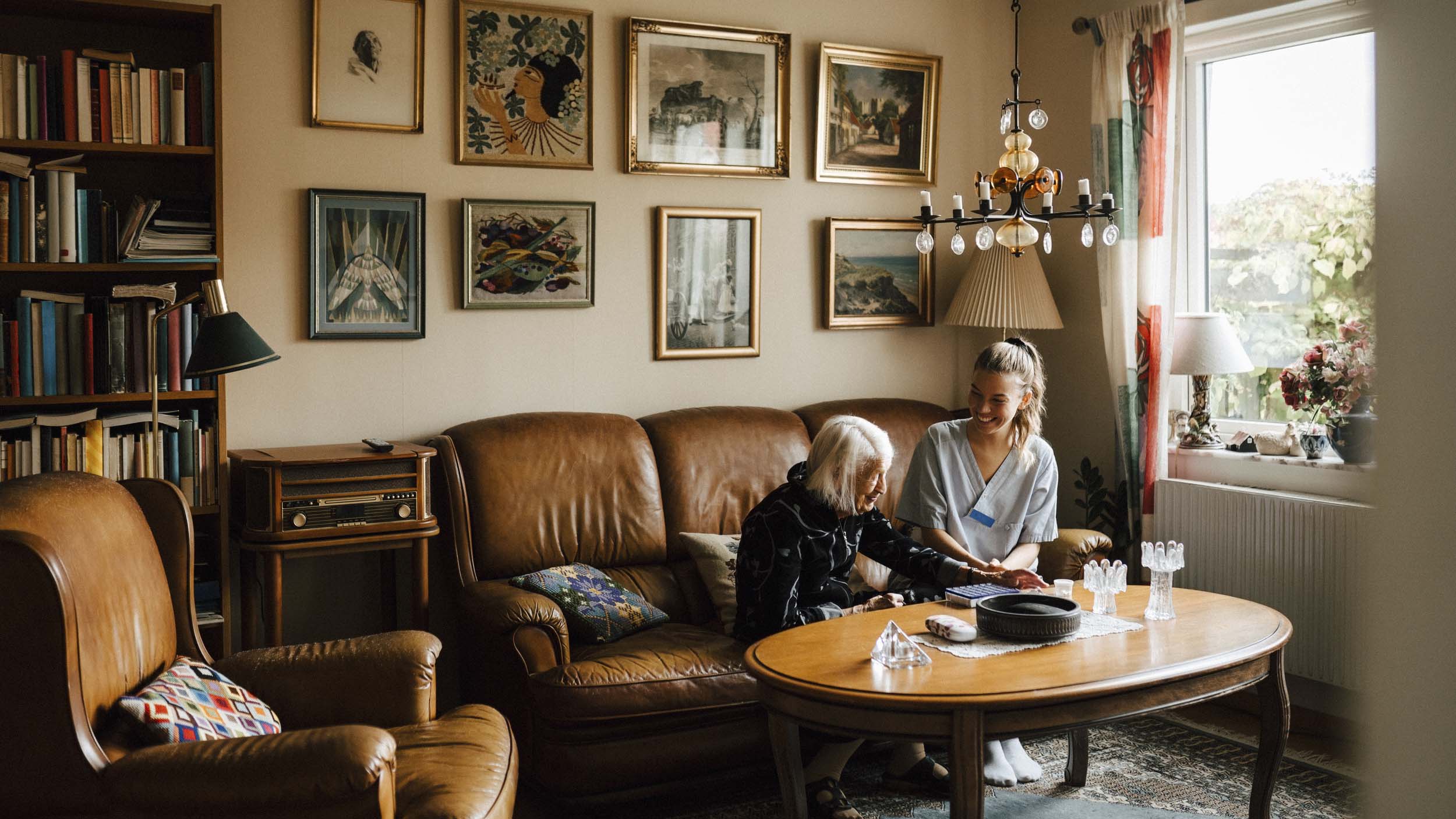 En äldre dam sitter i vardagsrumssoffan med en vårdare